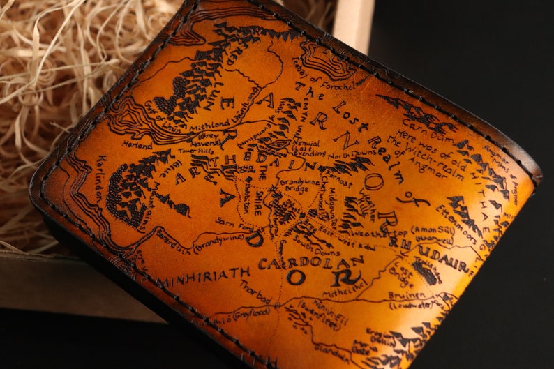 May include: A brown leather wallet with a map of Middle-earth from The Lord of the Rings engraved on the front. The map includes the Shire, Mordor, and the Misty Mountains.