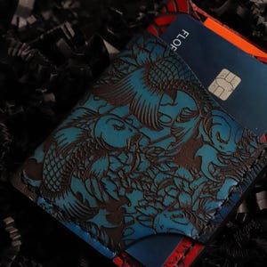 May include: A blue leather wallet with a koi fish design and a red and black border. The wallet has a card slot and is holding a blue credit card with the word "FLO" printed on it.
