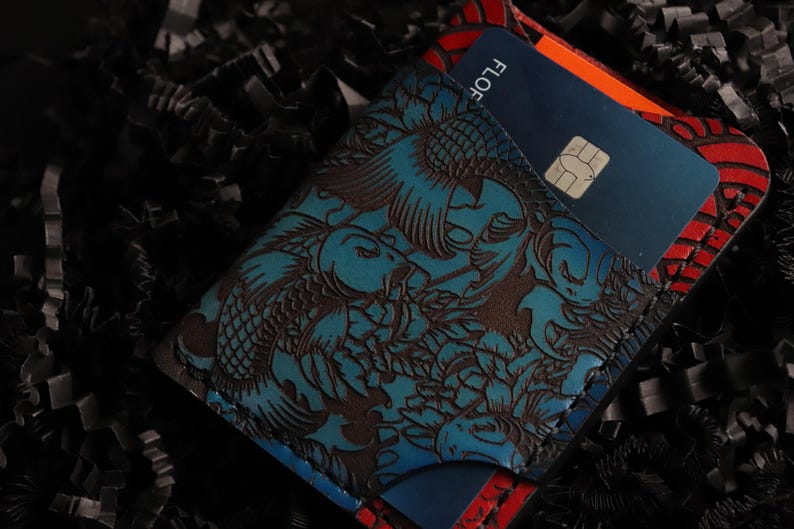 May include: A blue leather wallet with a koi fish design and a red and black border. The wallet has a card slot and is holding a blue credit card with the word "FLO" printed on it.