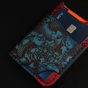 May include: A blue and red leather wallet with a koi fish design. The wallet has a slot for cards and a flap that closes with a snap.