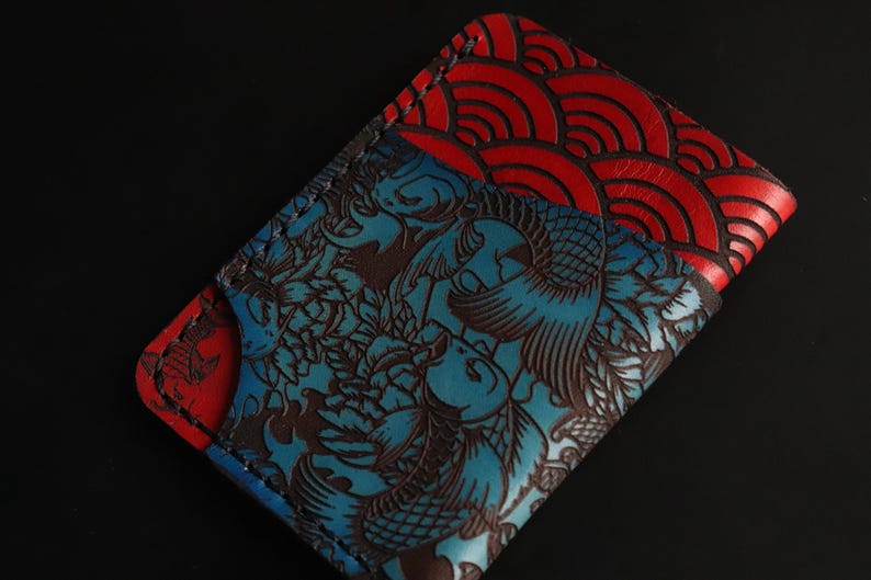 May include: A blue and red leather wallet with a Japanese-inspired design. One side features a blue background with a brown floral pattern and the other side features a red background with a wave pattern.