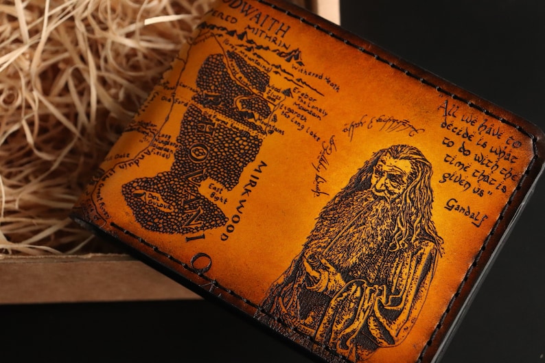 May include: A brown leather wallet with a map of Middle-earth from The Lord of the Rings. The map includes the text "Mirkwood" and "East Bight". The wallet also features a portrait of Gandalf the Grey with the quote "All we have to decide is what to do with the time that is given us."