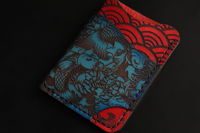May include: A blue and red leather wallet with a Japanese koi fish and wave pattern. The wallet is stitched with a dark brown thread.
