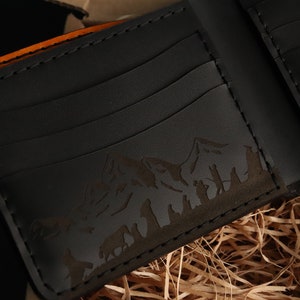 May include: A black leather wallet with an orange edge and an embossed mountain scene. The wallet has multiple card slots.