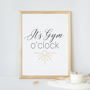 May include: A framed print with the text "It's Gym o'clock" in black cursive font and a gold sun graphic. The print is on a white background and is framed in a light wood frame.