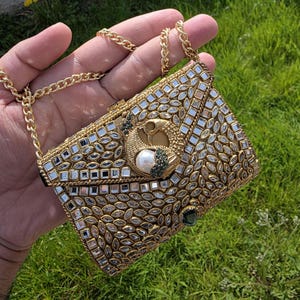May include: A gold clutch bag with a chain strap, adorned with mirrored squares and leaf-shaped details. The clasp showcases a peacock motif with a pearl and green accents. A small, rectangular green gem is visible.