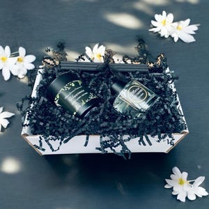 May include: A gift box containing a black candle labelled "Happiness", a clear perfume bottle, and black shredded paper. The box is decorated with white flowers and black incense sticks.
