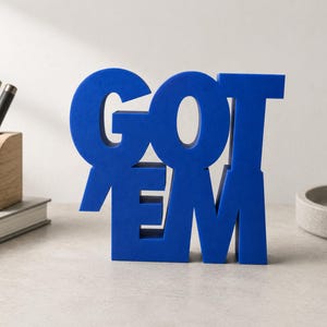 May include: A freestanding, bold blue sculptural sign featuring the text GOT EM in a stacked, block-letter format. The letters are constructed from a solid, matte-finish material, creating a three-dimensional display piece for a desk or shelf.
