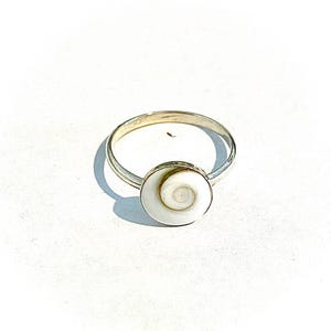 Puede incluir: Un anillo de plata con una incrustación de concha circular en espiral de color blanco y crema. La banda es delgada y pulida, sosteniendo la pieza de concha plana y redonda de forma segura en un engaste de metal. El diseño es simple y natural.