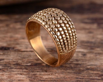 May include: A gold-colored ring with a textured surface of small, raised circles. The ring has a wide band and a domed top, creating a bold and eye-catching design. The ring is displayed on a wooden surface.