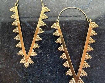 Peut inclure: Une paire de boucles d'oreilles créoles dorées avec un design en forme de V. Les boucles d'oreilles sont décorées de petites formes triangulaires.