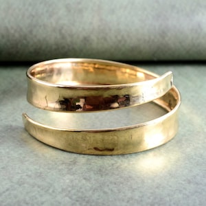 Puede incluir: Un brazalete de puño de metal martillado de color dorado con un diseño simple y moderno. El brazalete está hecho de dos piezas de metal que se unen en la parte posterior.