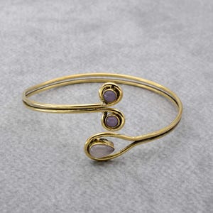 Pode incluir: Um bracelete dourado com um design único que apresenta três pedras de cor púrpura e branca. O bracelete é aberto e ajustável.