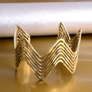 Op de afbeelding: Een goudkleurige manchetarmband met een gelaagd zigzagontwerp. De armband heeft een breed, open ontwerp en een gepolijste afwerking. De zigzags creëren een golfachtig patroon. De armband wordt gepresenteerd op een gouden oppervlak.