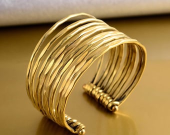 May include: A gold-colored cuff bracelet with a textured, hammered design. The bracelet features multiple parallel bands that curve around the wrist. The metal has a slightly aged appearance, giving it a vintage aesthetic. Jewelry.
