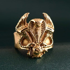 May include: A gold-toned ring featuring a detailed design of a cobra with wings and a motorcycle engine. The ring is a statement piece with intricate details.