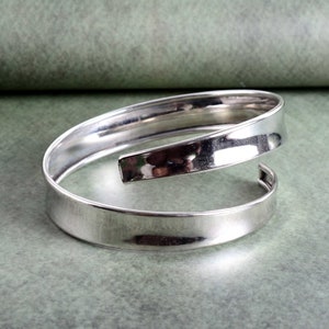 Könnte beinhalten: Ein silbernes Armreif-Armband mit einem einzigartigen Twist-Design. Das Armband besteht aus einem einzigen Stück Metall, das gebogen und verdreht ist, um einen einzigartigen und modernen Look zu schaffen.