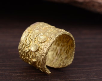 May include: A gold-toned ring with a textured surface. The ring is wide and has a unique, organic design.
