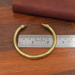 May include: A gold-coloured brass cuff bracelet with pointed ends. The bracelet is on a wooden surface next to a ruler. The text "MADE IN INDIA" is visible. The bracelet's diameter is approximately 6.35 cm.