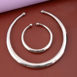 May include: Two silver bangles, one larger than the other, with rounded edges. The bangles are on a brown surface.