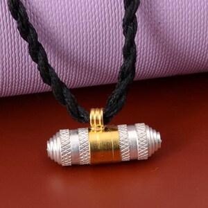 May include: A silver and gold cylindrical pendant with a braided black cord necklace. The pendant has a textured design and is suspended from a gold loop.