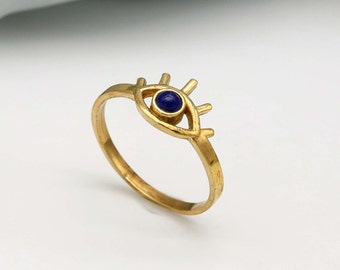 May include: A gold-colored ring featuring an eye design. The eye has a dark blue center stone and stylized lashes. The ring is set against a white background, highlighting the intricate details of the jewelry.