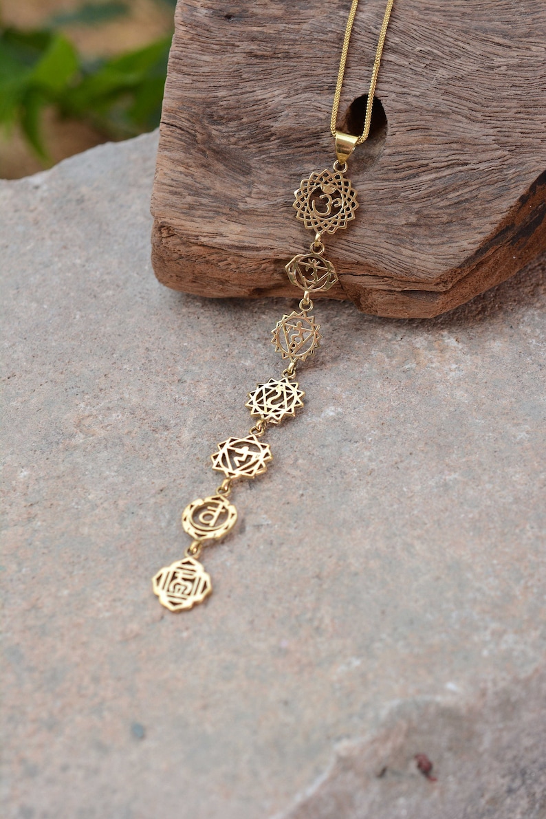 May include: A gold necklace with seven chakra symbols, including Om, hanging from a chain. The necklace is displayed on a wooden surface.
