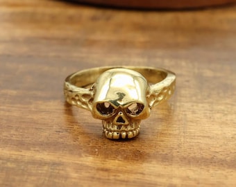 May include: A gold-coloured skull ring. The ring features a detailed skull design with eye sockets, a nose cavity, and teeth. The band has an ornate pattern. The ring is displayed on a wooden surface.
