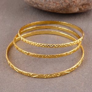 May include: Three gold-toned bangles with intricate etched designs. The bangles are stacked on top of each other.