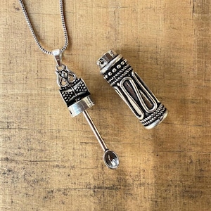 May include: A silver-colored pendant necklace with a decorative cylindrical container and a small spoon. The container is open, revealing the spoon. The necklace and container are on a wooden surface.
