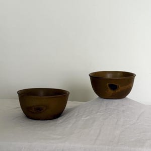 May include: Two round, deep bowls with a matte, dark brown finish. Each bowl features a distinct, darker, irregular organic-shaped mark on its exterior surface. The bowls have a smooth, curved rim and a simple, rounded base design.
