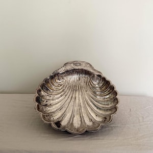 May include: A silver-toned metal dish shaped like a large scallop shell. The bowl features deep, radiating fluted grooves and a scalloped edge, with a polished finish that shows signs of natural tarnish and patina throughout the surface.
