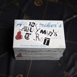Op de afbeelding: Een witte doos met de tekst "Alley Striker's The Alleyman's Tarot" en een rode peerillustratie. De doos is gelabeld met "A proud American Company" en "Alley Striker's Two Stick Marbles".