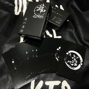 Puede incluir: Una caja negra con caracteres japoneses blancos que se leen "花 の 言葉" que se traduce como "Lenguaje de las flores". La caja est&aacute; abierta y contiene una baraja de cartas de juego negras con dise&ntilde;os blancos. Las cartas tienen un c&iacute;rculo con caracteres japoneses en el centro.
