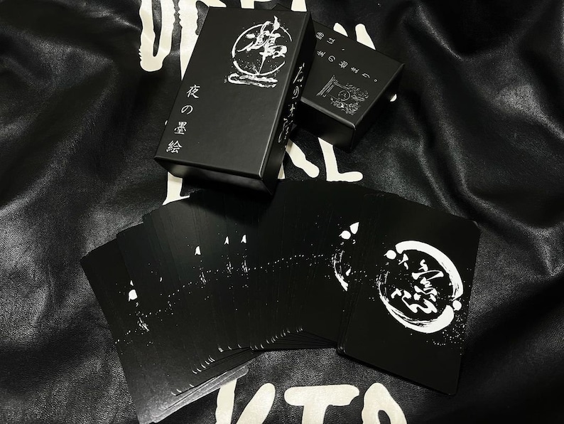 Puede incluir: Una caja negra con caracteres japoneses blancos que se leen "花 の 言葉" que se traduce como "Lenguaje de las flores". La caja est&aacute; abierta y contiene una baraja de cartas de juego negras con dise&ntilde;os blancos. Las cartas tienen un c&iacute;rculo con caracteres japoneses en el centro.