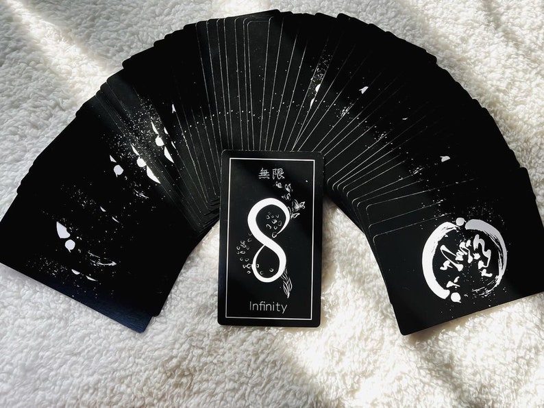 Puede incluir: Una baraja de cartas de tarot negras con tinta blanca, que presenta un s&iacute;mbolo de infinito y la palabra "Infinity" en la carta de la parte delantera. Las cartas est&aacute;n desplegadas en semic&iacute;rculo.