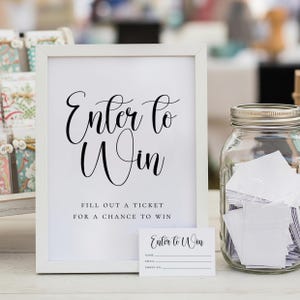 May include: A white framed sign with the words "Enter to Win" in elegant script. Below, it reads "Fill out a ticket for a chance to win." A small card and a glass jar filled with tickets are also present.