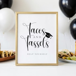 Op de afbeelding: Een ingelijst bord met de woorden "Tacos and Tassels" in elegant schrift, vergezeld van een afstudeerpet-afbeelding. De zin "Help Yourself" staat eronder gedrukt. Het bord staat in een gouden lijst, met zwarte en witte ballonnen op de achtergrond.