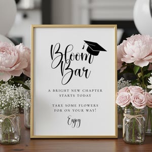 Op de afbeelding: Een ingelijst bord met de woorden "Bloom Bar" in elegant schrift, bekroond met een illustratie van een afstudeerpet. Het bord bevat de zinnen "A Bright New Chapter Starts Today" en "Take Some Flowers For On Your Way! Enjoy!"