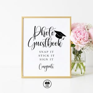 May include: A framed sign with gold trim reads "Photo Guestbook" in elegant script, with a graduation cap illustration. Below, it says "Snap it, Stick it, Sign it, Congrats." A bouquet of pink flowers sits nearby.