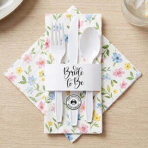 May include: A white paper napkin featuring a watercolor floral pattern in pink, blue, and yellow, bundled with a white plastic fork, knife, and spoon. A white paper band wraps around the set, printed with the black text Bride to Be.