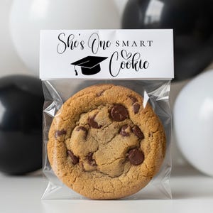 Op de afbeelding: Een chocolate chip cookie in een doorzichtige plastic zak met een wit label met de tekst "She's One Smart Cookie" met een afstudeerpet-afbeelding. Zwarte en witte ballonnen op de achtergrond.