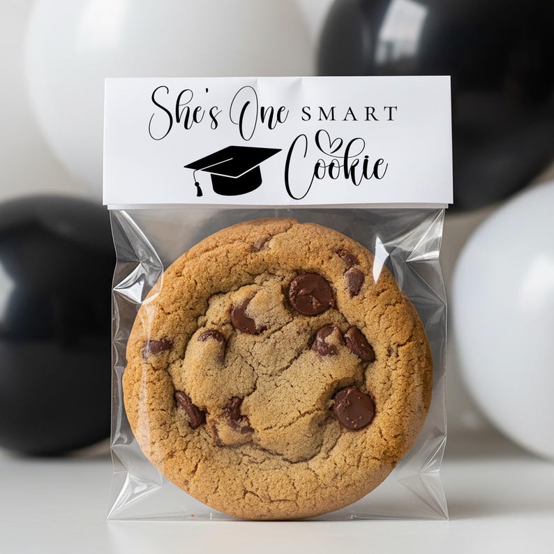 Op de afbeelding: Een chocolate chip cookie in een doorzichtige plastic zak met een wit label met de tekst "She's One Smart Cookie" met een afstudeerpet-afbeelding. Zwarte en witte ballonnen op de achtergrond.