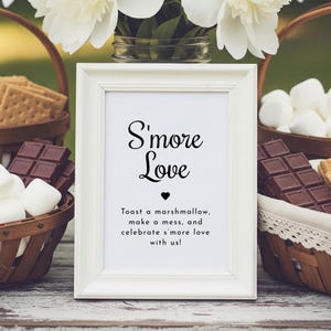 May include: A white framed sign with the text "S'more Love" and a heart symbol. The sign is surrounded by baskets of marshmallows, chocolate bars, and graham crackers, suggesting a s'mores theme. White flowers are in the background.