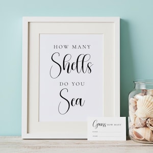 May include: A white framed sign with black text that reads HOW MANY Shells DO YOU Sea, accompanied by a small matching guess card and a clear glass jar filled with various seashells, all displayed on a light wooden surface.