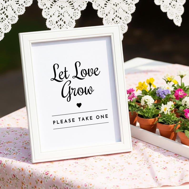 May include: A white framed sign with the text "Let Love Grow" in black script, a heart symbol, and the words "PLEASE TAKE ONE". The sign is on a floral patterned tablecloth, with small potted flowers in the background.