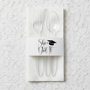 Op de afbeelding: Een set van doorzichtig plastic bestek, bestaande uit een vork, mes en lepel, gepresenteerd op een witte servet. Een witte band met de tekst "She Did It" en een afstudeerhoedje omwikkelt het bestek. Geschikt voor een afstudeerfeest.