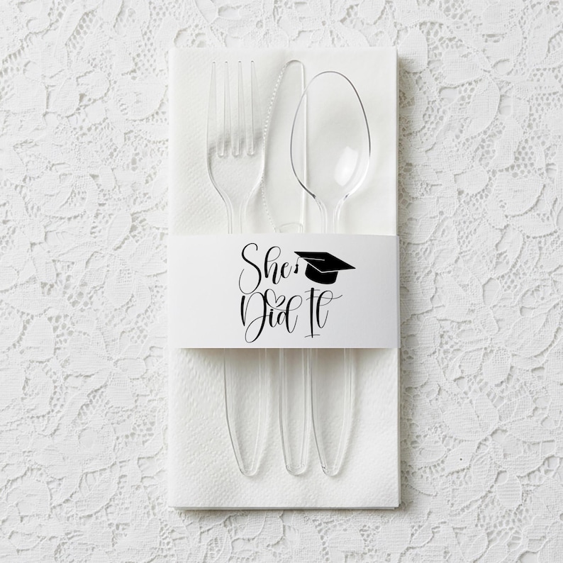 Op de afbeelding: Een set van doorzichtig plastic bestek, bestaande uit een vork, mes en lepel, gepresenteerd op een witte servet. Een witte band met de tekst "She Did It" en een afstudeerhoedje omwikkelt het bestek. Geschikt voor een afstudeerfeest.