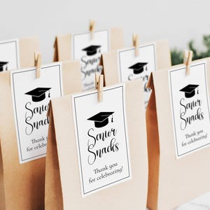 Op de afbeelding: Verschillende bruine papieren zakken met witte labels met de tekst "Senior Snacks" en een afbeelding van een afstudeerhoed. Elke zak is vastgezet met een kleine houten knijper. De tekst "Thank you for celebrating!" is ook aanwezig.