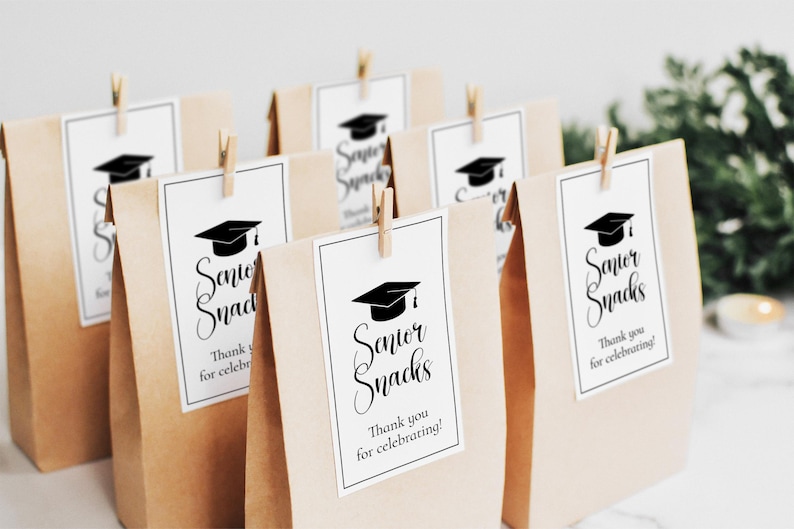 Op de afbeelding: Verschillende bruine papieren zakken met witte labels met de tekst "Senior Snacks" en een afbeelding van een afstudeerhoed. Elke zak is vastgezet met een kleine houten knijper. De tekst "Thank you for celebrating!" is ook aanwezig.